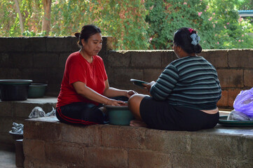 Two women sit indoors facing each other, opening the tray and using a plastic plate to take peanuts from the basin.