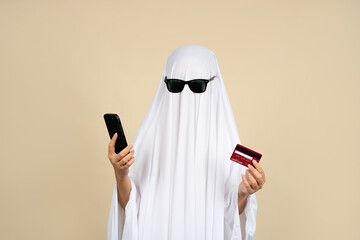 Person in white ghost costume holding a credit card and smartphone on a beige background. Celebrating Halloween.