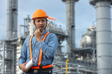 male engineer holding blueprints and using a walkie-talkie at industrial plant, representing construction management, energy industry, and occupational safety.