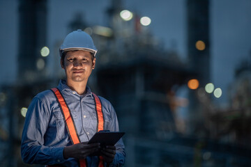Confident industrial engineer wearing safety helmet and reflective vest holding digital tablet during night shift at oil refinery or power plant, focusing on smart facility management and safety.