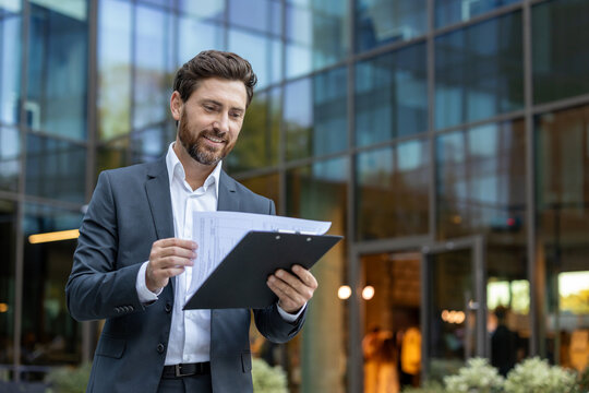 Confident businessman reviewing important papers on a clipboard with a content smile, standing in front of a modern glass office building, symbolizing success and corporate life - Powered by Adobe