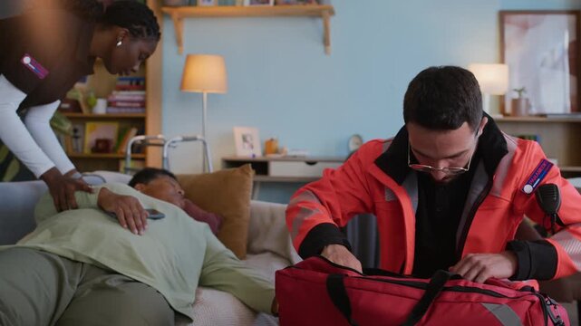 Handheld shot of two rescuers in paramedic uniform rushing to provide first aid to senior unconscious woman lying on sofa