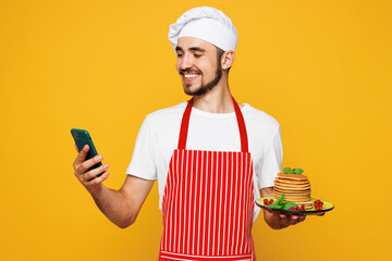 Young housewife housekeeper chef cook baker man wear red apron toque hat hold stack of pancakes use mobile cell phone isolated on plain yellow orange background studio portrait. Cooking food concept.