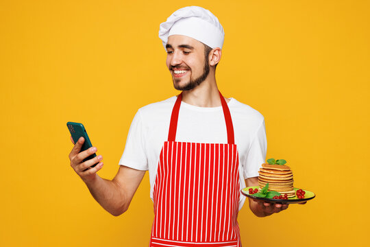 Young housewife housekeeper chef cook baker man wear red apron toque hat hold stack of pancakes use mobile cell phone isolated on plain yellow orange background studio portrait. Cooking food concept. - Powered by Adobe