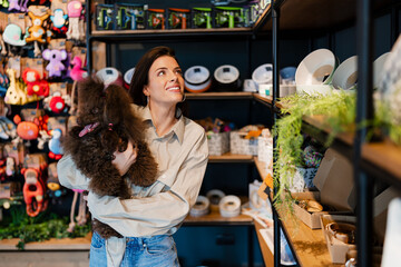 Woman smiling and holding her poodle dog while looking at colorful pet harnesses and supplies in a retail pet shop, conceptualizing pet ownership and care