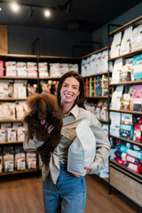 Woman smiling and holding her fluffy brown poodle and a bag of pet food while shopping in a modern...
