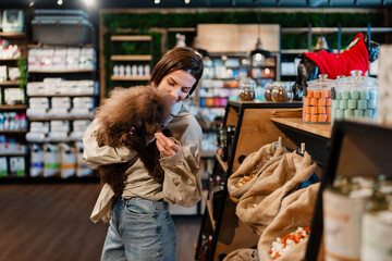 Woman holding a cute brown mini poodle and selecting high-quality pet food and treats in a modern...
