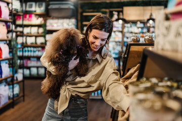 Woman holding a cute brown mini poodle and selecting high-quality pet food and treats in a modern...