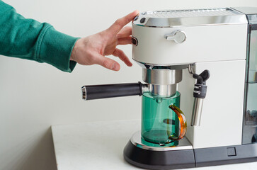 Cozy morning with mans hands pressing coffee machine button for espresso into green glass cup at home