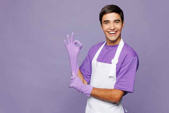 Side view young smiling happy satisfied man wear violet t-shirt apron while doing housework tidy up show ok okay gesture isolated on plain pastel light purple background studio. Housekeeping concept.