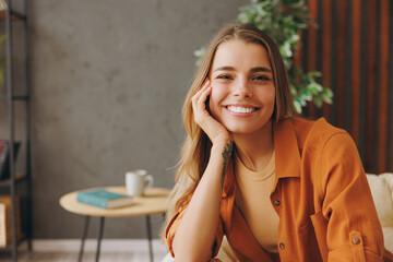 Close up young smiling happy woman she wears orange casual clothes sits in chair looking camera stay at home hotel flat rest relax spend free spare time in living room indoor. People lounge concept.