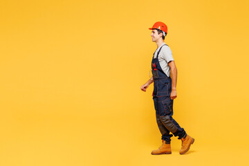 Full body side profile view young happy employee laborer handyman man wear helmet overall walk go isolated on plain yellow background. Instruments for renovation apartment room. Repair home concept.
