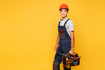 Side view young employee laborer handyman man wear helmet overall hold in hand toolbox isolated on plain yellow background. Instrument accessories for renovation apartment room. Repair home concept.