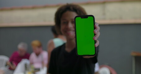 Teenager holding smartphone with green screen, smiling confidently, outdoors at a casual gathering with diverse adults interacting in the blurred background
