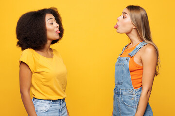 Side profile view two young funny friends buddies women wear denim casual clothes together show tongue look to each other isolated on plain yellow orange background studio portrait. Lifestyle concept. © ViDi Studio