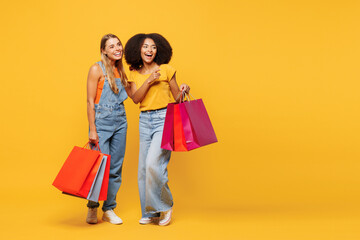 Full body two young fun friends women wear casual clothes hold shopping package bags together point index finer aside on area isolated on plain yellow orange background. Black Friday buy day concept.