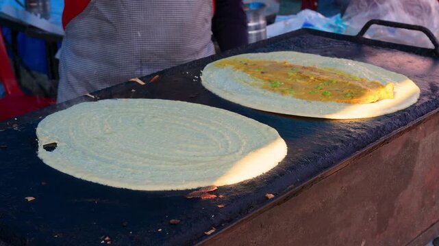 Close-up video of a street vendor skillfully spreading dosa batter on a hot griddle, capturing the sizzling sound and rustic charm of rural India's outdoor food culture.