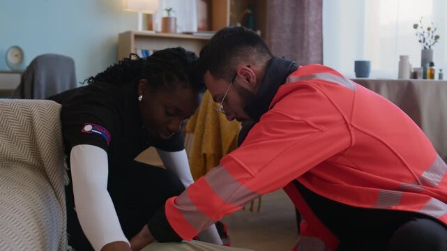 Handheld shot of two diverse healthcare workers in uniform checking vital signs of senior unresponsive woman lying on floor in flat