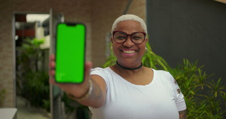 African American woman extending smartphone forward, green screen Chroma Key visible, confident smile, blurred background, emphasizing digital interface and connection outdoors