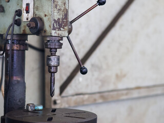 Close-up of a vintage, industrial-style, vertical drill press, showing the chuck, drill bit, and feed handles against a blurred workshop background.