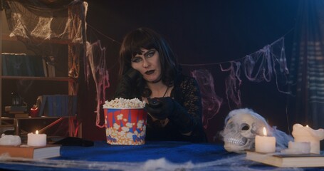 Women dressed as witch eat popcorn and watch a boring movie on TV in a dimly lit, festive Halloween...