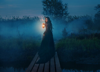 art photo real person silhouette gothic woman hand hold lantern lamp walking across bridge pier over river dense fog mist night nature dark blue sky lake. black dress. Scared fantasy girl looking back