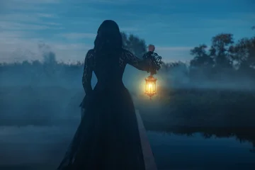 photo real person silhouette gothic woman hand hold lantern lamp walking across bridge over river dense fog mist night nature dark dramatic blue sky lake. long black dress back rear view. Fantasy girl © kharchenkoirina