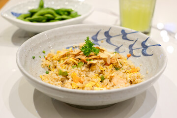 Asian fried rice served in a ceramic bowl with fried shallots and parsley on top, accompanied by edamame and green drink in the background. Perfect for restaurant menu design, Asian cuisine