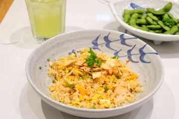 Asian fried rice served in a ceramic bowl with fried shallots and parsley on top, accompanied by edamame and green drink in the background. Perfect for restaurant menu design, Asian cuisine