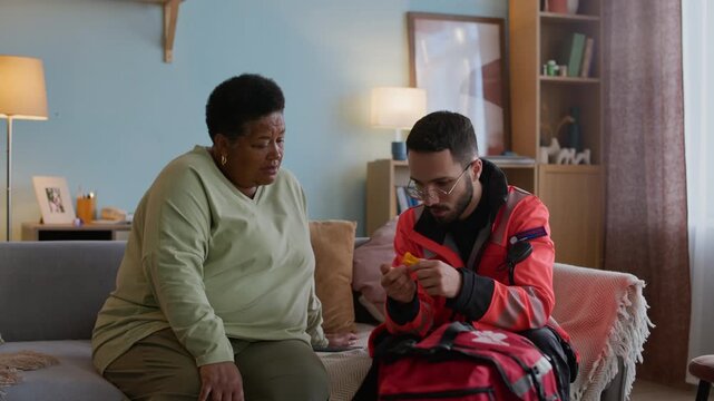 Medium long shot of young male multiethnic paramedic in uniform giving pills to senior African American woman sitting on sofa during emergency at home