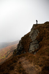 Fototapeta premium Hiker stands on rocky outcrop overlooking misty mountain landscape in autumn. Autumn hiking in Carpathian Mountains, Ukraine