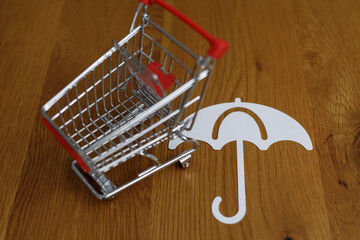 Conceptual shot of a shopping cart and umbrella on a wooden surface. Represents protection,...