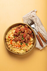 Traditional dish of Maghreb cuisine, a Moroccan couscous with lamb or beef, vegetables, chickpeas, and peas in a rustic bowl with cutlery on yellow background