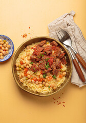 Traditional dish of Maghreb cuisine, a Moroccan couscous with lamb or beef, vegetables, chickpeas, and peas in a rustic bowl with cutlery on yellow background