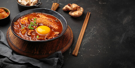 Top view of spicy kimchi jjigae soup in a black bowl with egg yolk, enoki, shiitake, tofu and green onions, with chopsticks and cooking ingredients, cuisine of Korea, copy space