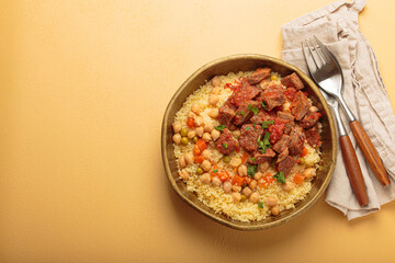 Traditional dish of Maghreb cuisine, a Moroccan couscous with lamb or beef, vegetables, chickpeas, and peas in a rustic bowl with cutlery on yellow background, copy space