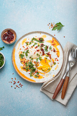 Turkish eggs cilbir, a delicious and colorful breakfast with poached eggs on yogurt, seasoned with spices and fresh herbs, arranged on a blue background