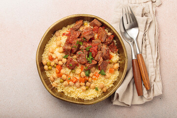 Traditional dish of Maghreb cuisine, a Moroccan couscous with lamb or beef, vegetables, chickpeas, and peas in a rustic bowl with cutlery on beige background