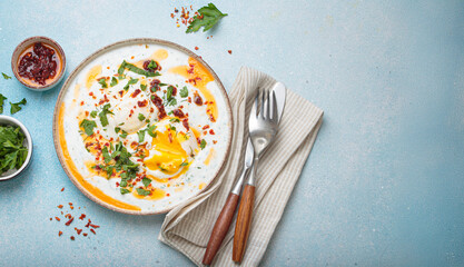 Turkish eggs cilbir, a delicious and colorful breakfast with poached eggs on yogurt, seasoned with spices and fresh herbs, arranged on a blue background, copy space