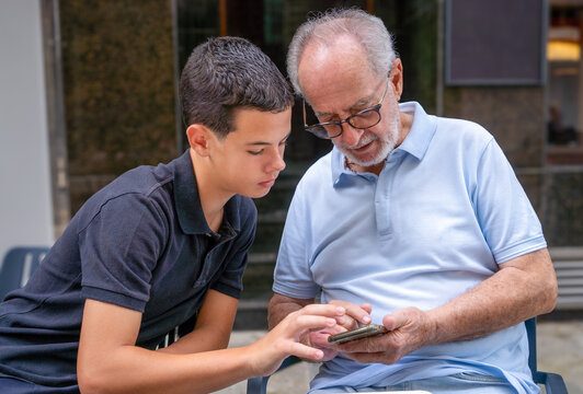 Grandson helping his grandfather to using the smartphone, sitting on a terrace outdoors. Technology concept.