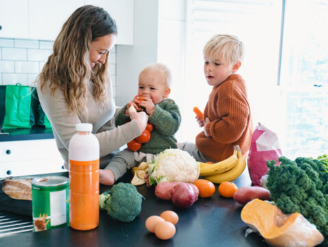 Mum holding Baby boy taking a bite on a bag of tomato while older brother looking at him