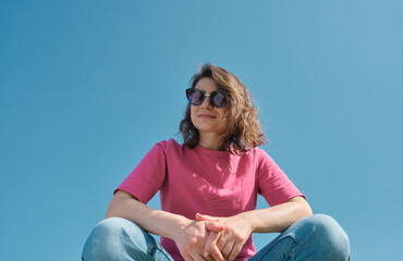 Beautiful carefree young woman in sunglasses and pink t-shirt sitting and looking down at camera on blue sky background low angle shot. Copy space
