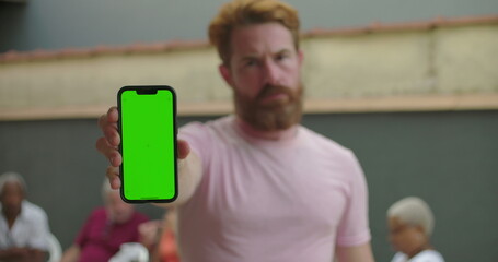 Man holding smartphone with green screen at arm’s length, serious expression, standing outdoors, people blurred in background, customizable display focus