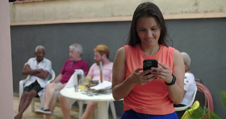 Woman smiling softly while looking at smartphone screen, standing outdoors, casual group conversation in blurred background, enjoying digital content
