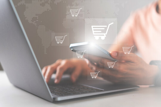 Hand using smartphone and laptop for online shopping, with shopping cart icons overlay, represents e-commerce, digital marketing, and global retail technology trends.