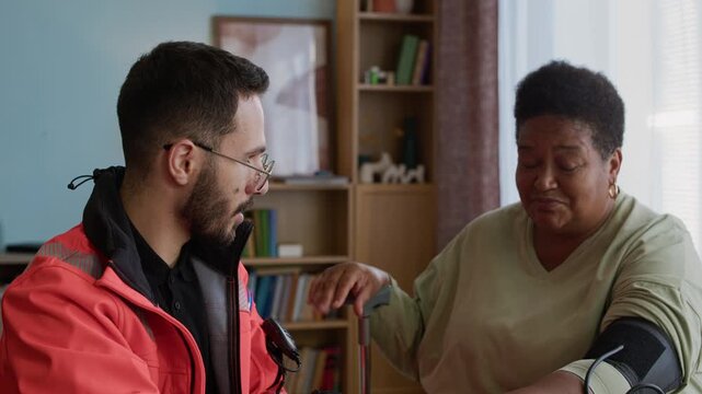 Zoom out shot of male specialist in orange paramedic uniform talking to senior woman measuring blood pressure at home