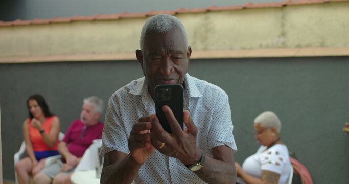 African American Elderly man smiling while looking at smartphone screen, standing outdoors with casual group conversation in blurred background, engaged with digital interaction - Powered by Adobe