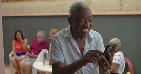 African American Elderly man smiling and using a smartphone outdoors, engaged and joyful, with a group of diverse adults sitting in the background, social gathering atmosphere