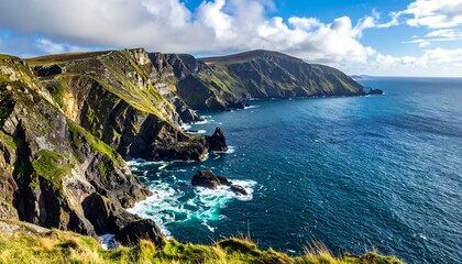 Fototapeta premium A scenic coastal view of a rugged, green cliffside meeting the vibrant blue ocean under a partly cloudy sky. Waves crash against the rocky shore