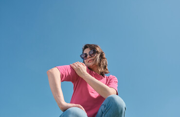 Beautiful carefree young woman in sunglasses and pink t-shirt smiling and looking down at camera on blue sky background low angle shot. Copy space